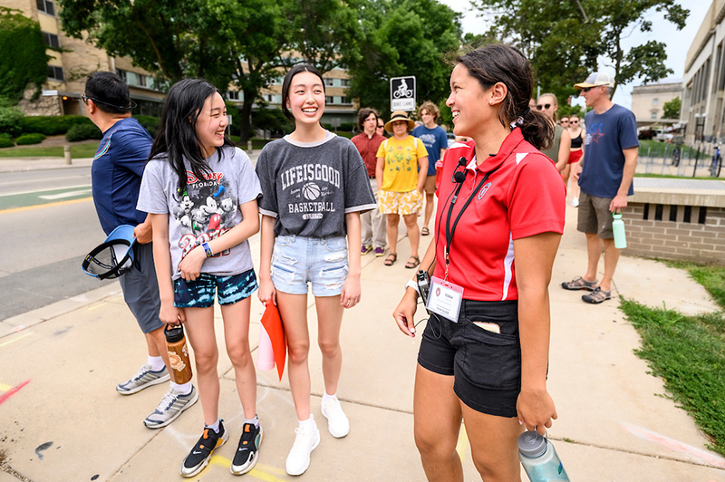 A student in a red shirt with a lanyard smiles and talks to two children on the sidewalk.