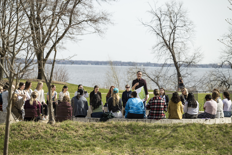 An adult speaks to a group of students seated in a circle in front of a lake.